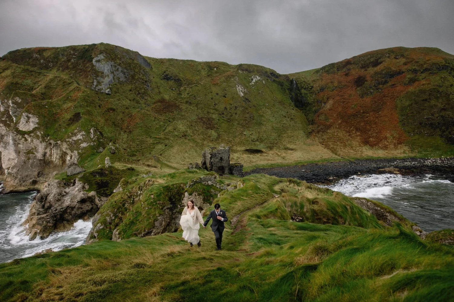A bride and groom walking hand-in-hand along a grassy coastline with rocky terrain, green hills, a river, and overcast sky in the background.