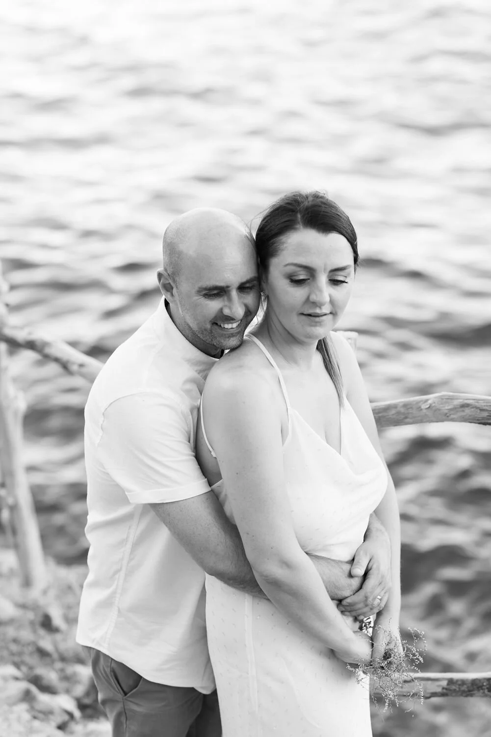 A couple embraces by a body of water, with the man smiling and the woman looking contemplative, in a black and white photograph.