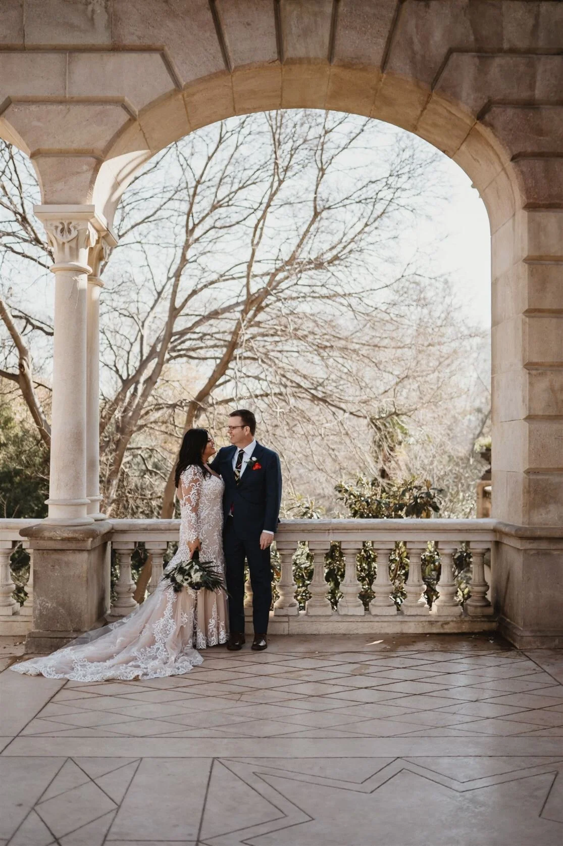 A bride and groom in wedding attire standing on a stone balcony with decorative balustrades, under a stone archway, with leafless trees in the background.