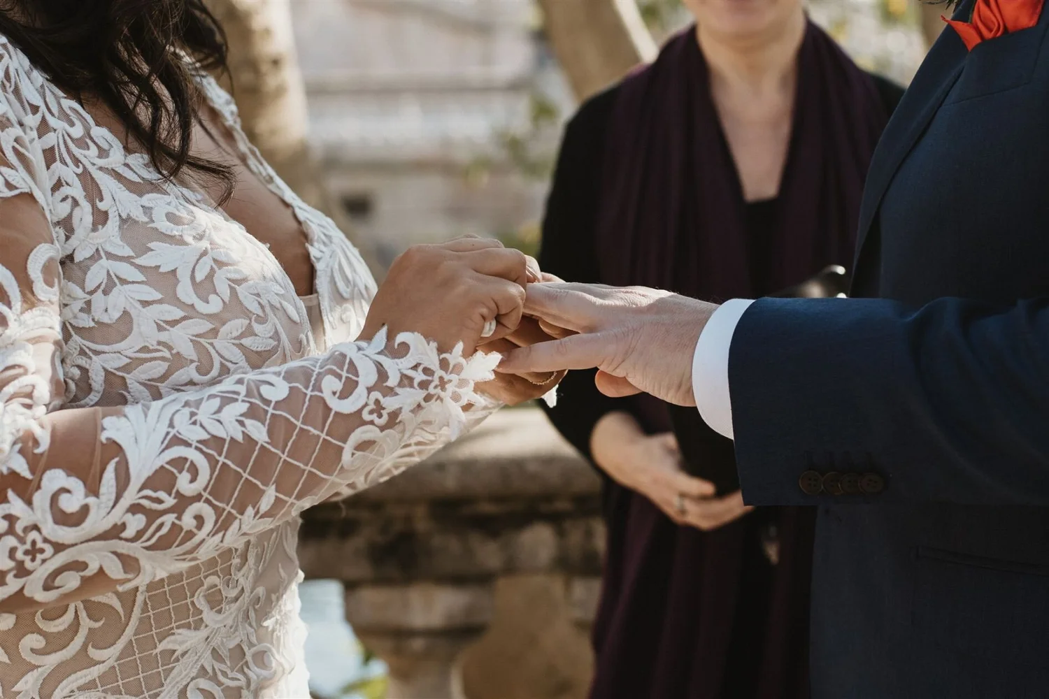 A couple exchanging wedding rings during an outdoor wedding ceremony, with a woman in a white lace dress and a man in a navy suit, and an officiant standing behind them.