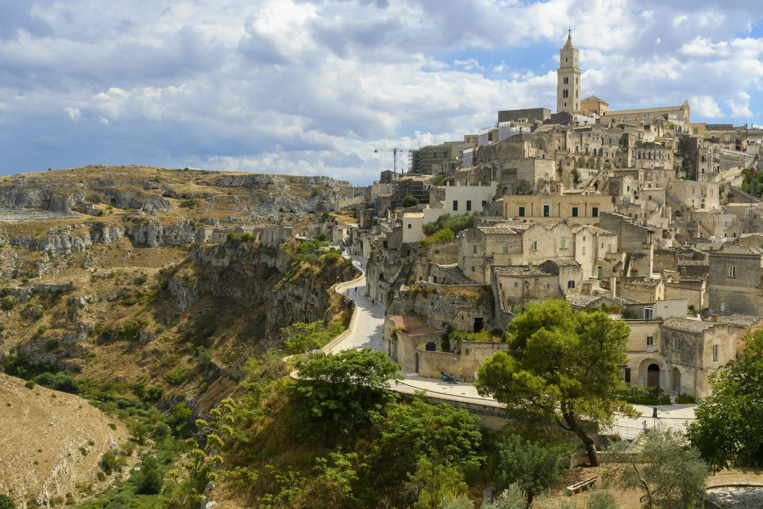 A hilltop town with stone buildings and a tall church tower, overlooking a landscape with rocky terrain and sparse vegetation under a partly cloudy sky.