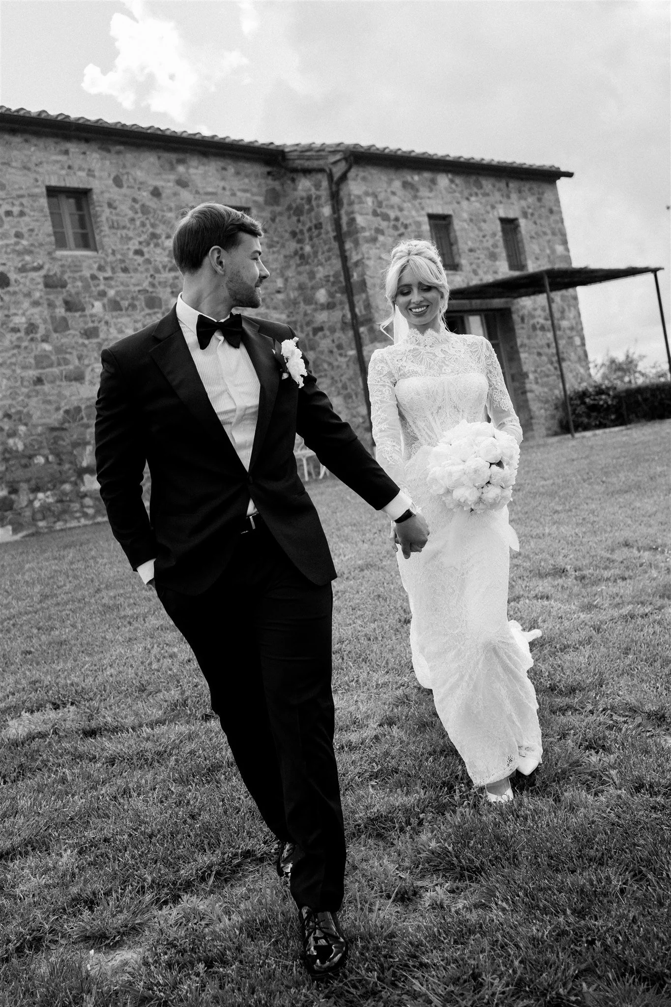 A black and white photo of a bride and groom walking outdoors. The groom is smiling and looking at the bride, dressed in a tuxedo with a bow tie. The bride, wearing a lace wedding dress and holding a bouquet, is smiling down and holding hands with the groom. They are in front of a brick building with a cloudy sky above.