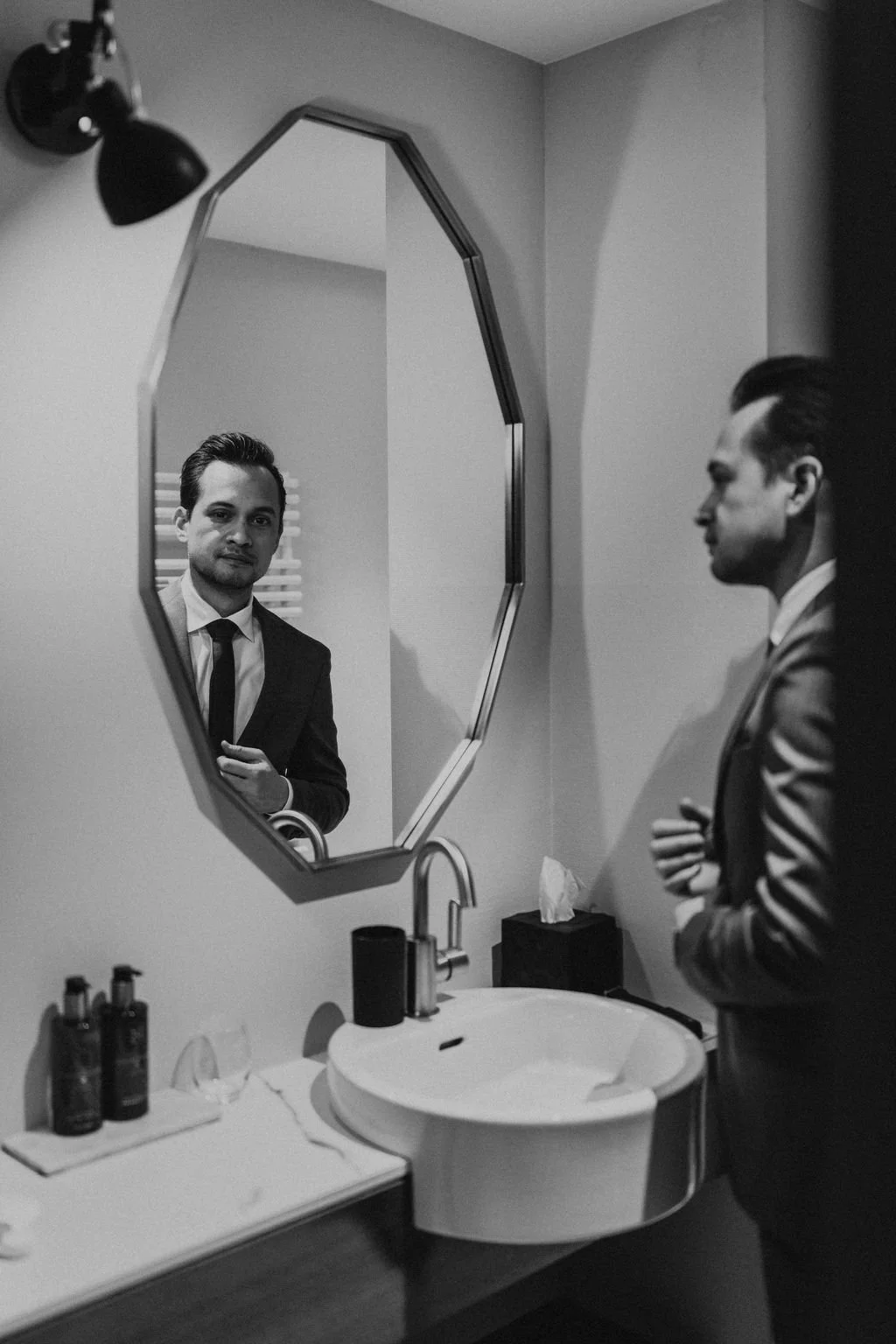 A man in a suit looking at himself in a bathroom mirror, with toiletries on the sink counter and a tissue box nearby.