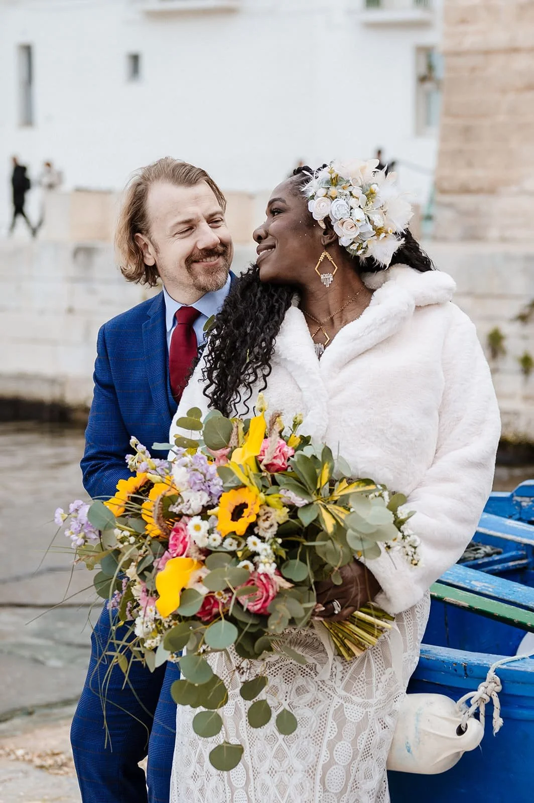 A couple at a wedding, the woman holding a colorful bouquet of flowers, both smiling and looking at each other, with a white building and water in the background.