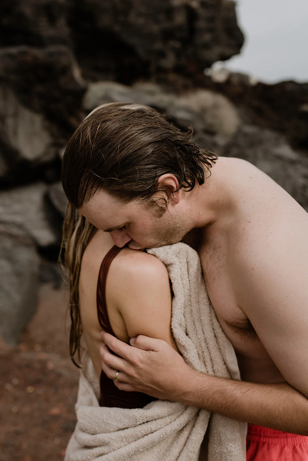 A man and woman embrace outdoors, with the man kissing the woman's shoulder as she holds him gently, surrounded by rocky terrain.