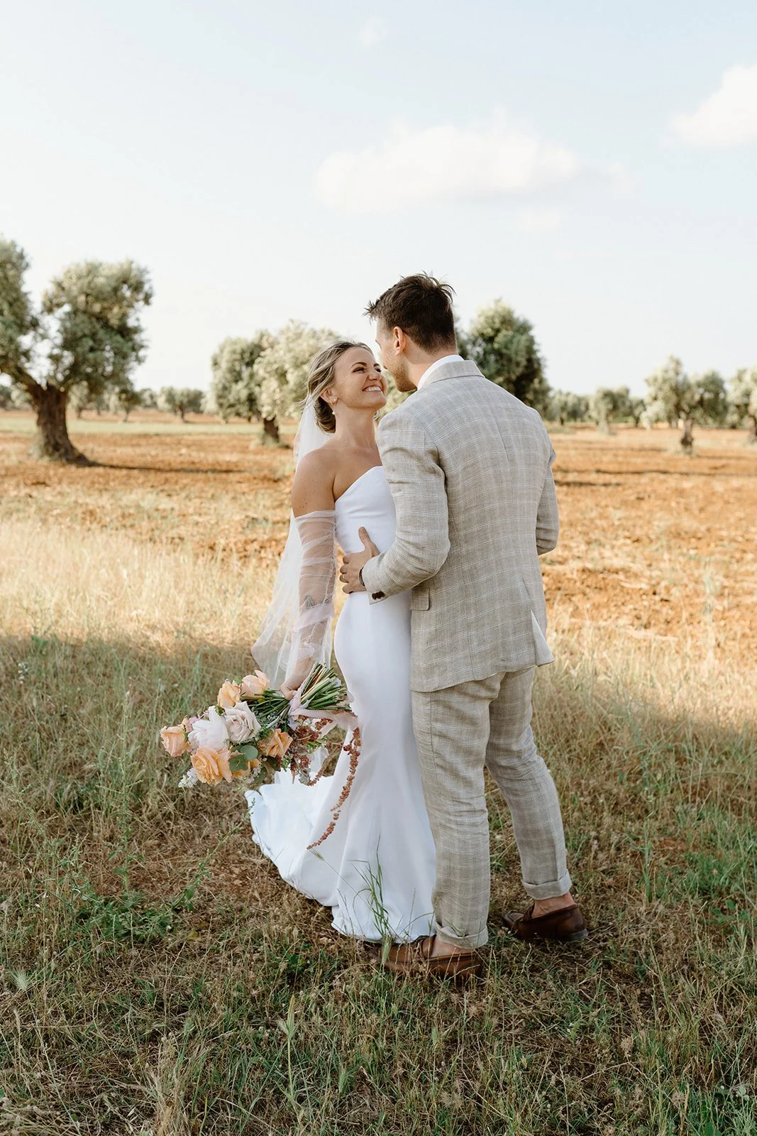 A bride and groom share a joyful moment in an outdoor setting with trees and a partly cloudy sky; the bride holds a bouquet of pastel flowers.