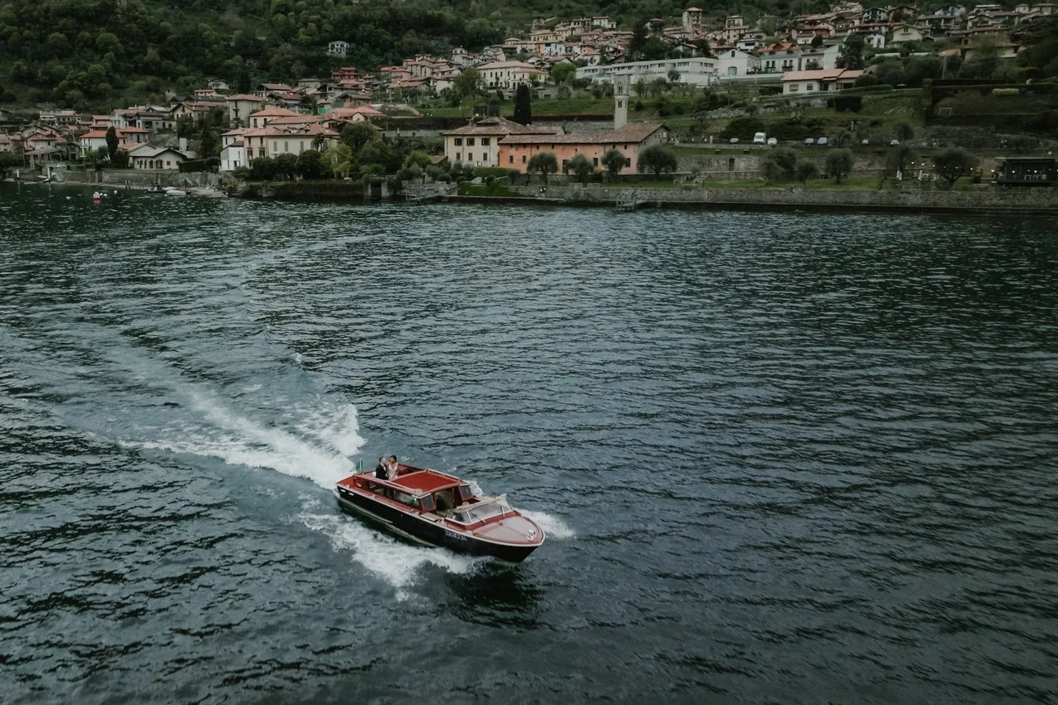 A boat with two people on board traveling across a body of water near a European hillside town with traditional houses and lush green trees.