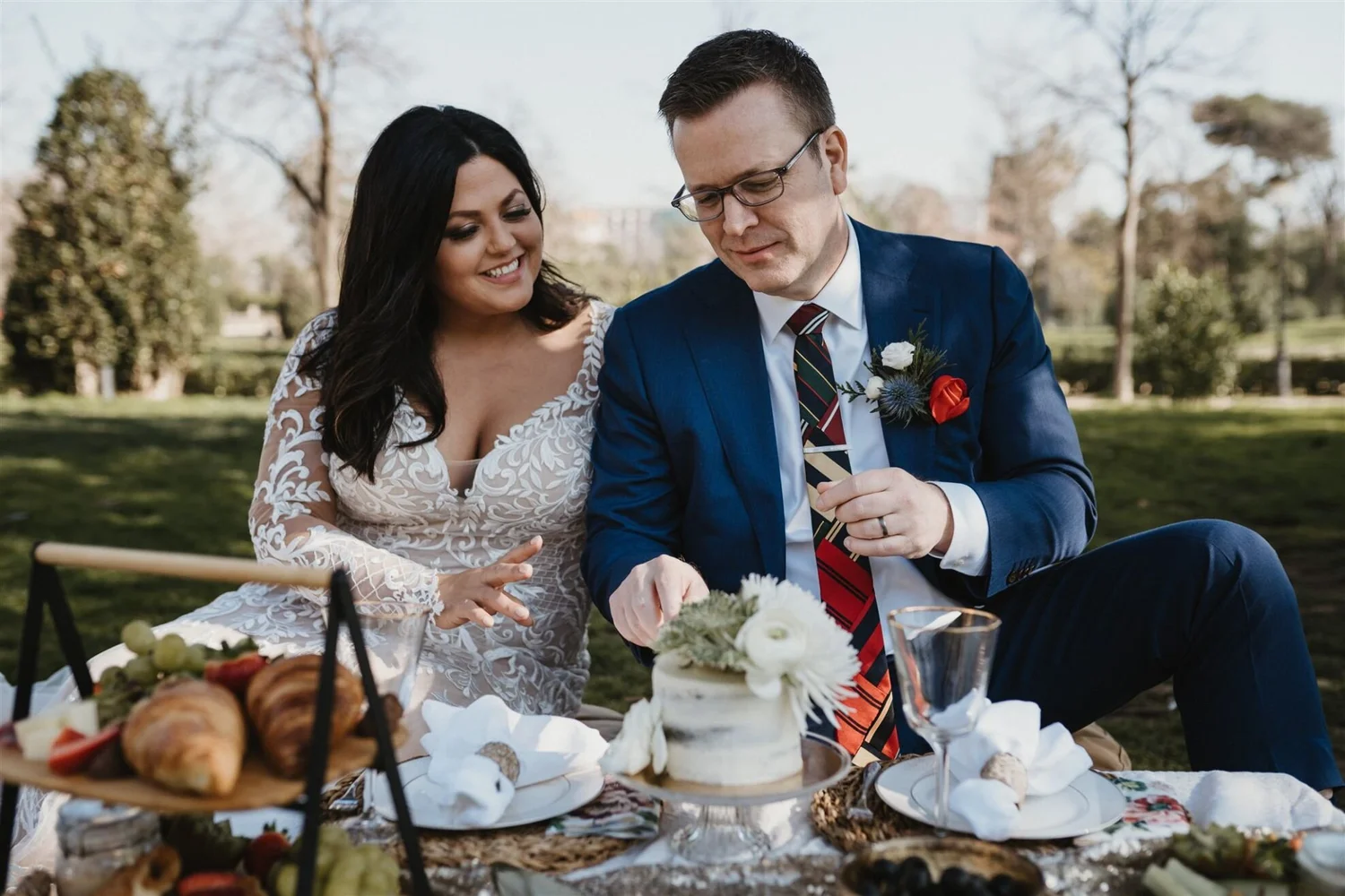 A bride and groom sitting outdoors at a wedding reception, smiling and looking at each other. The bride has dark hair and is wearing a white lace dress. The groom is wearing glasses, a blue suit, and a striped tie, with a boutonniere. There is a tabl