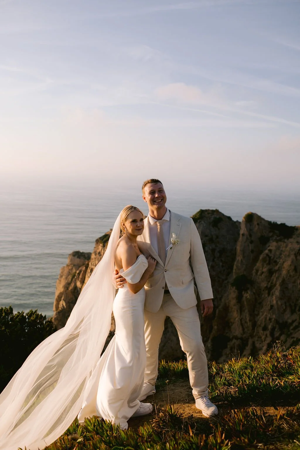A bride and groom in wedding attire standing on a cliffside overlooking the ocean, smiling at the camera during sunset.