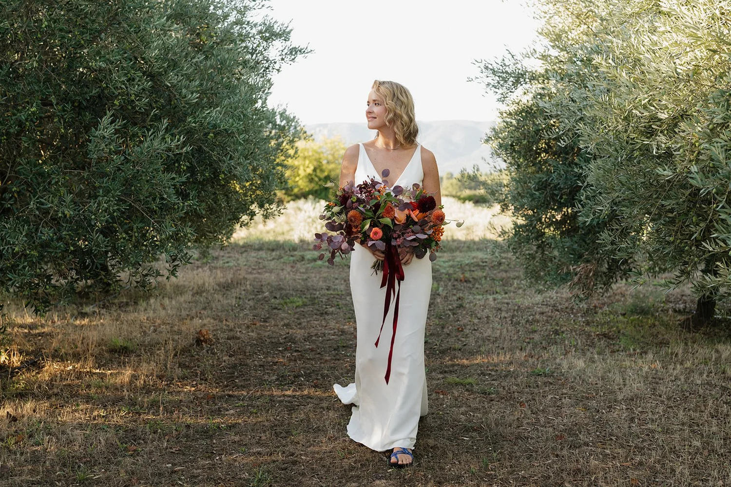 A woman in a white dress walking through a grove of olive trees while holding a large bouquet of colorful flowers.