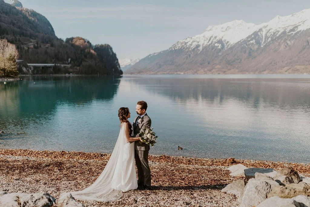 A bride and groom standing by a lake, holding hands, with snow-capped mountains in the background.