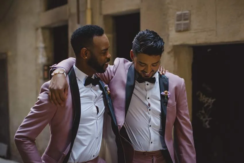 Two men in pink tuxedos and black bowties sharing a moment, smiling and with their arms around each other's shoulders.