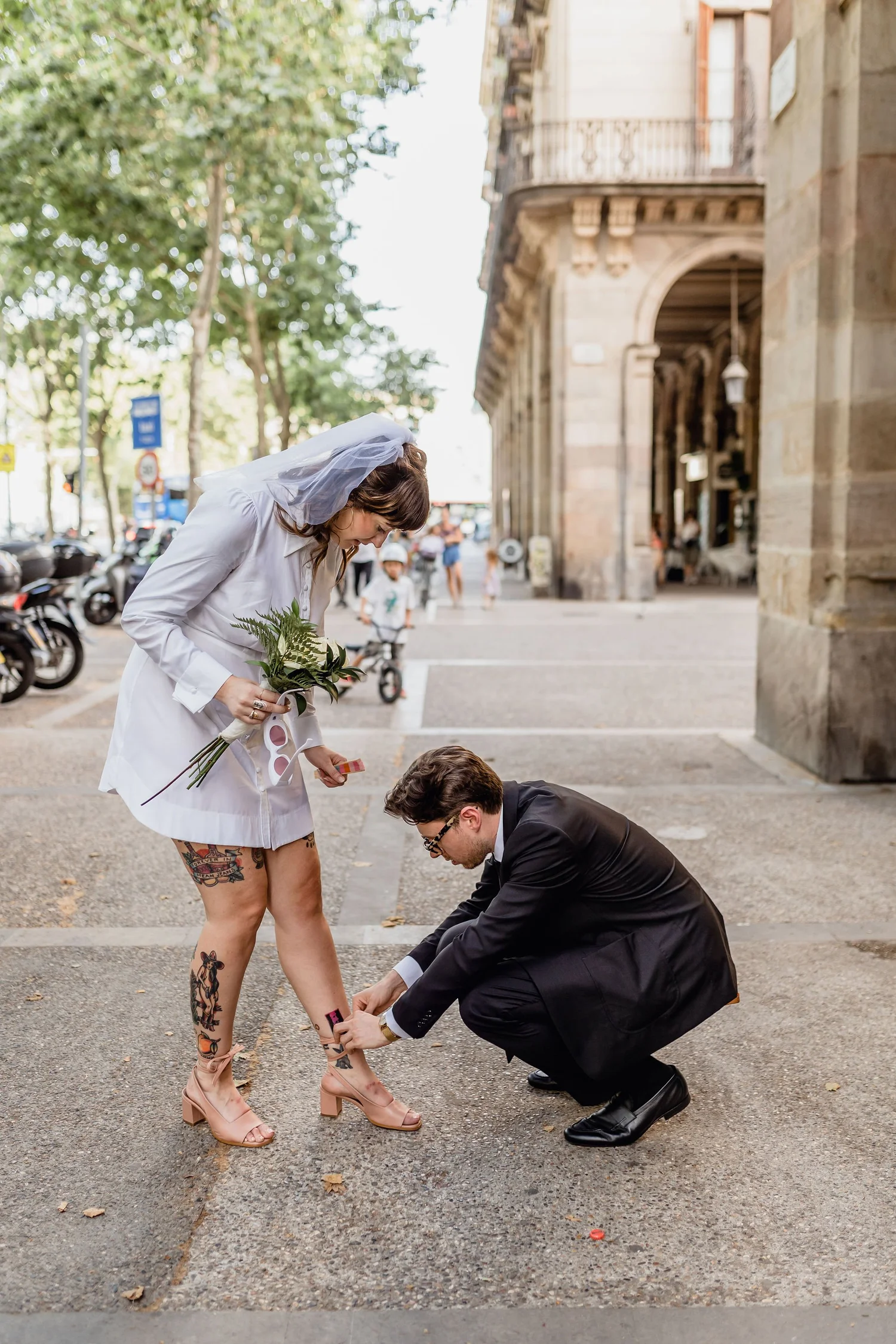 A bride with tattoos and wearing a white dress and veil is being pinned with a boutonniere by a man in a suit on the street in front of a historic building.