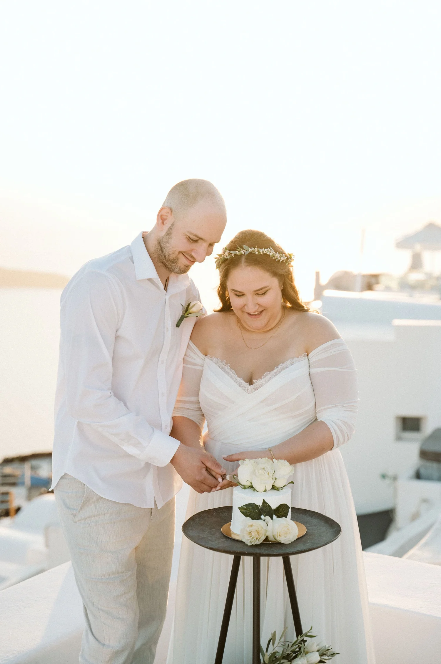A couple in wedding attire cutting a wedding cake outdoors during sunset. The bride is wearing a white dress and a floral crown, and the groom is in a white shirt and light-colored pants. The wedding cake is decorated with white roses and greenery, p