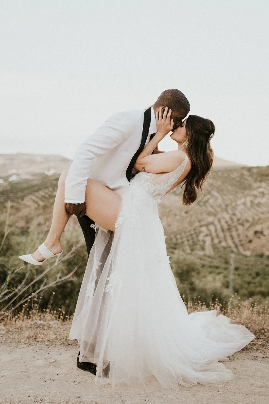 A bride and groom share a kiss outdoors, with the groom carrying the bride. The bride is wearing a white wedding gown and heels, and the groom is in a white suit with a black tie. The background features rolling hills and a cloudy sky.