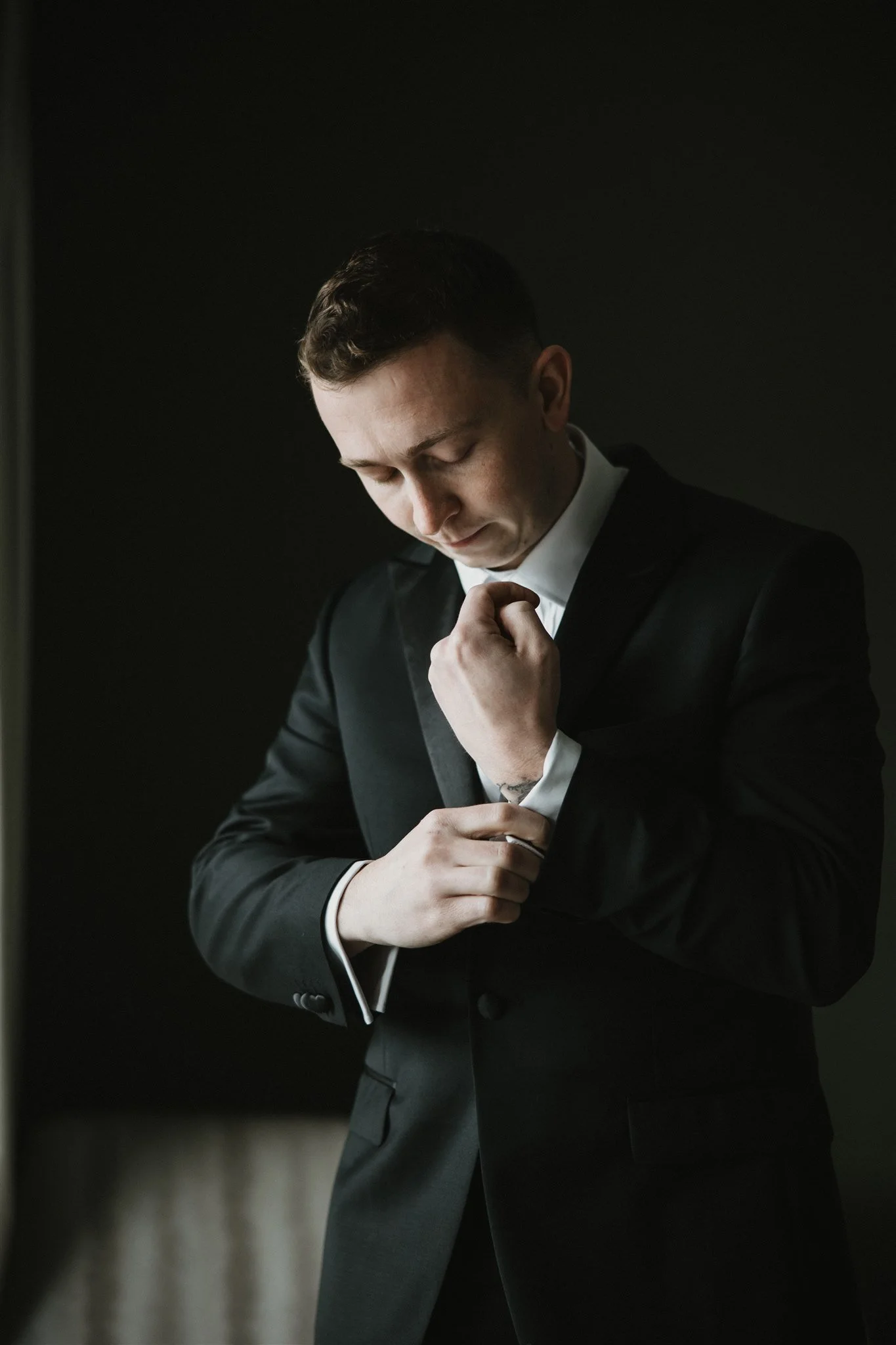 A young man in a black suit and white shirt adjusting his cufflinks, standing against a dark background.