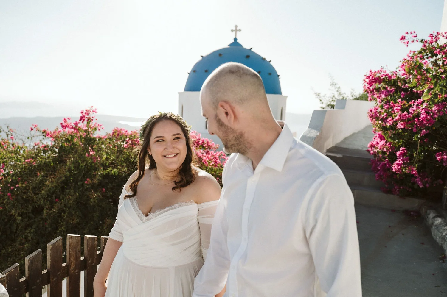 A smiling woman and man dressed in white, standing outdoors with pink flowering bushes and a small church with a blue dome and cross in the background.