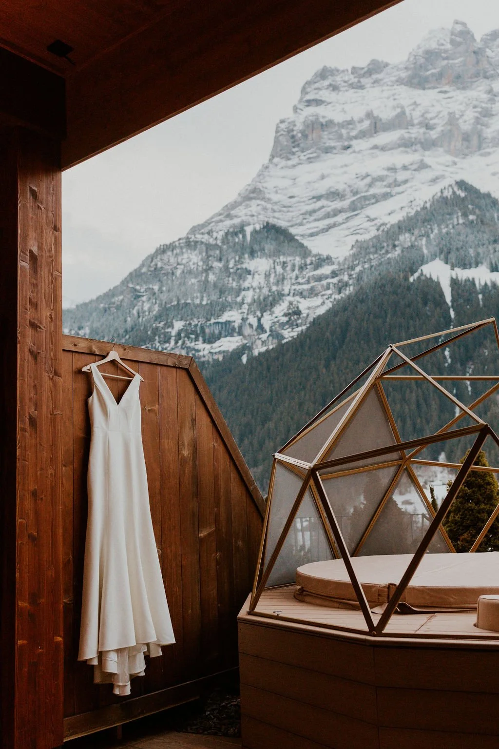 A white wedding dress hanging on a wooden wall with mountains in the background, and a hot tub with a glass enclosure on a wooden deck.