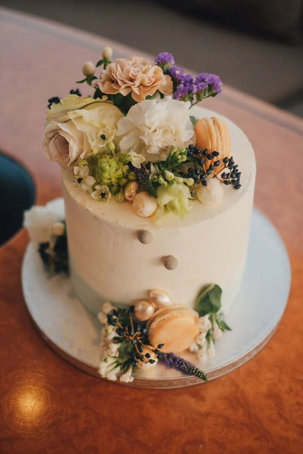 A white frosted cake decorated with assorted flowers, berries, and macarons, sitting on a silver cake board on a wooden surface.