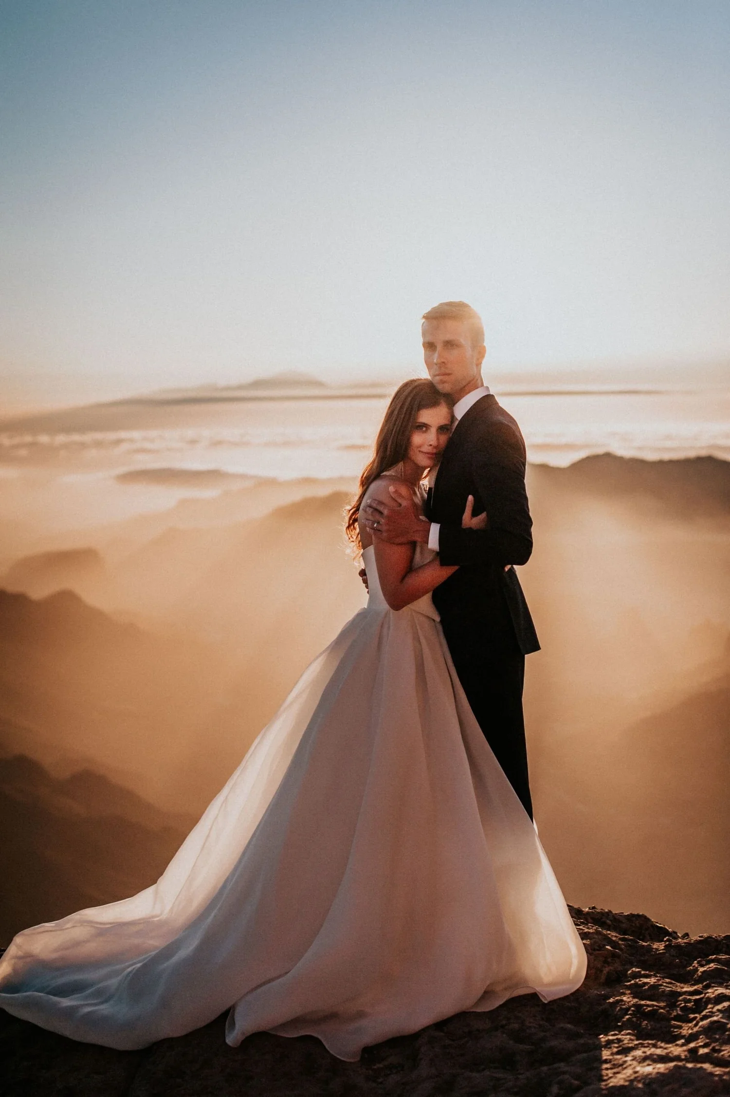 A couple dressed in formal wedding attire standing on a rocky beach at sunset, with the woman in a white gown and the man in a black tuxedo, embracing each other.