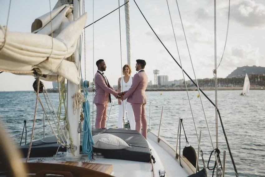 Two men in pink suits holding hands and facing each other on a boat, with a woman in a white dress standing behind them, in a harbor with sailboats and a city skyline in the background.