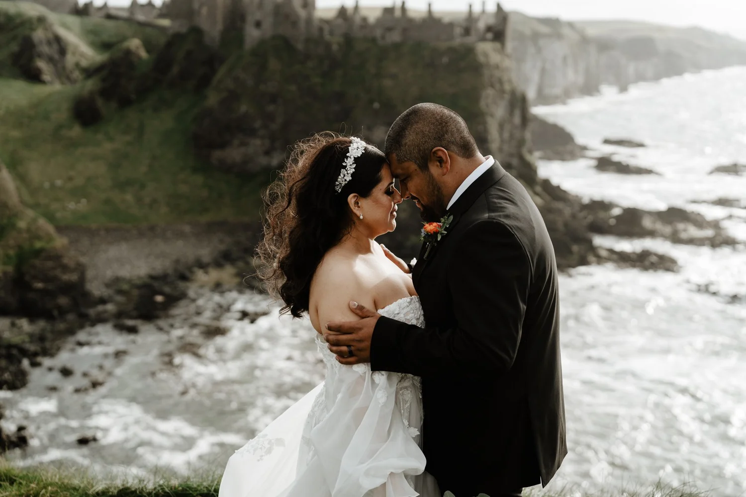 A couple dressed in wedding attire embracing on a grassy area near the ocean with cliffs in the background.
