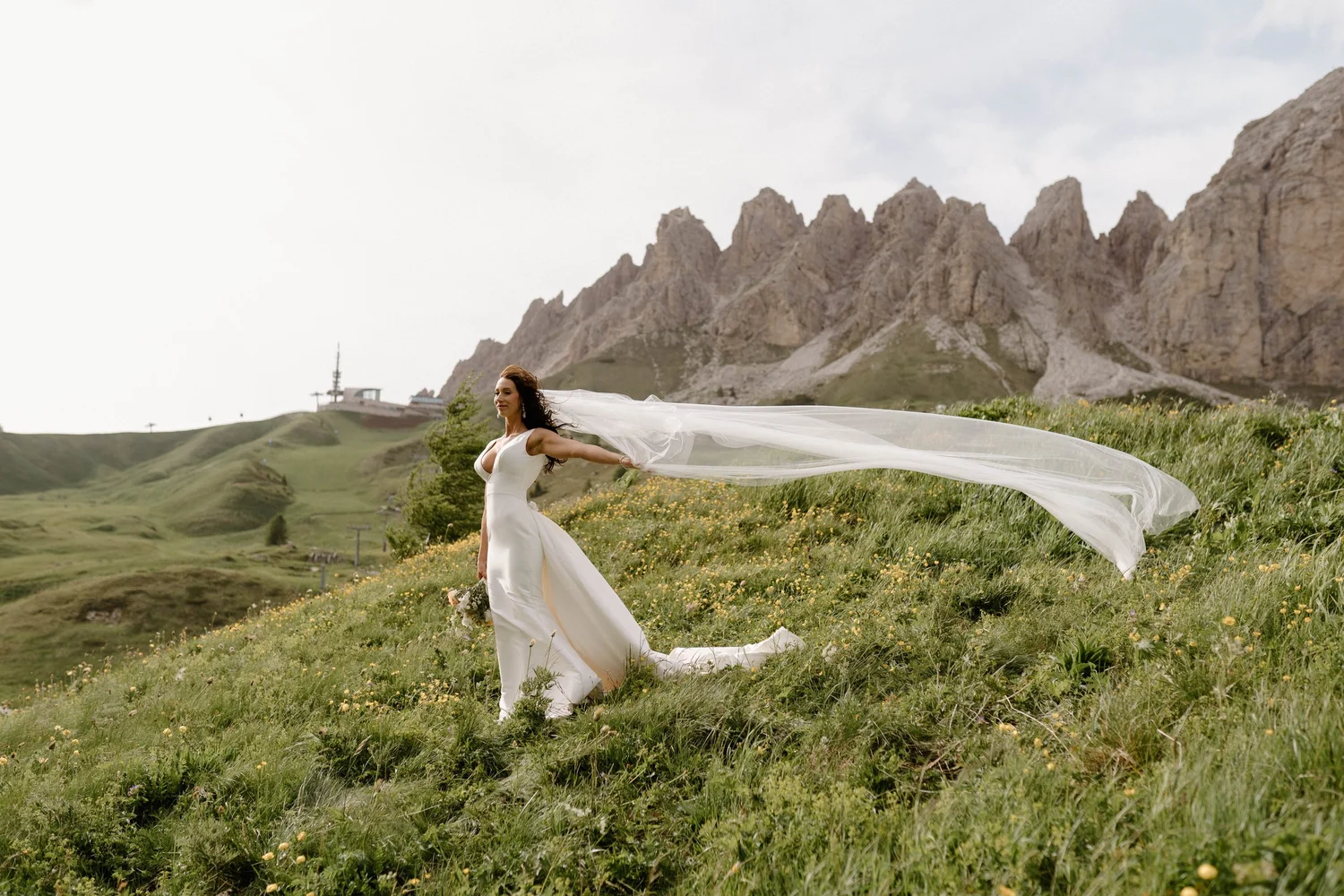A bride in a white wedding dress with a long veil walking through a grassy field with mountains in the background.