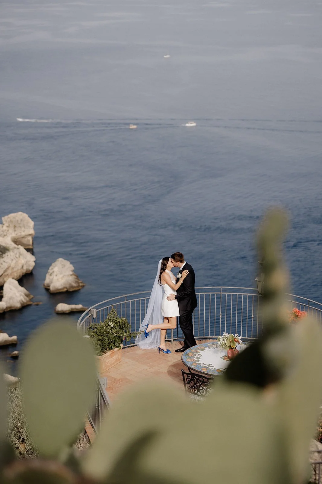 A couple dressed in wedding attire sharing a kiss on a seaside terrace, with boats in the water in the background.