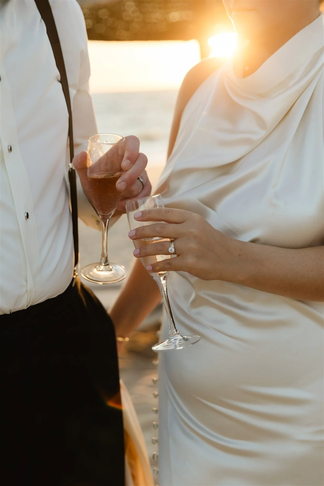 Close-up of a couple holding champagne glasses during sunset at a beach wedding, with the ocean in the background.