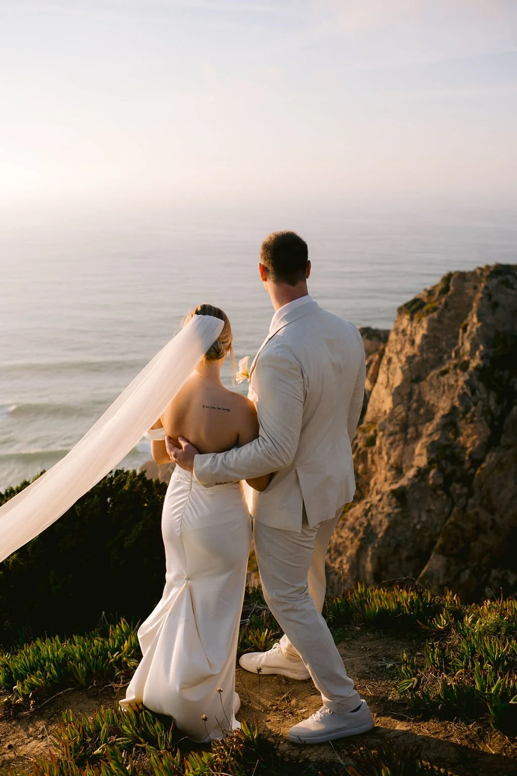 A bride and groom standing on a cliff overlooking the ocean at sunset.