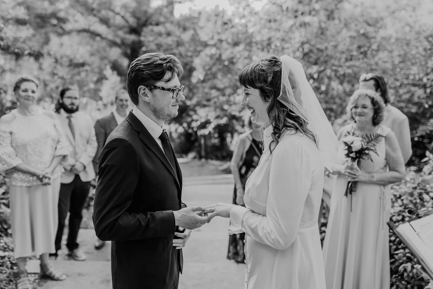 A black and white photo of a wedding ceremony outdoors with a couple holding hands and looking into each other's eyes. The groom is wearing glasses and a tuxedo, and the bride is in a white dress with a veil. Bridesmaids and groomsmen are standing in