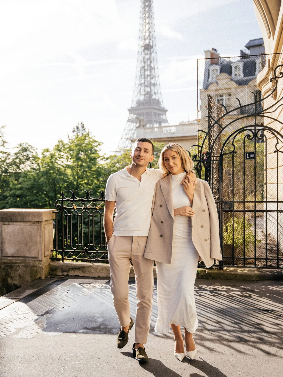 A couple walking together outdoors, near the Eiffel Tower in Paris, France, with greenery and decorative fencing in the background.