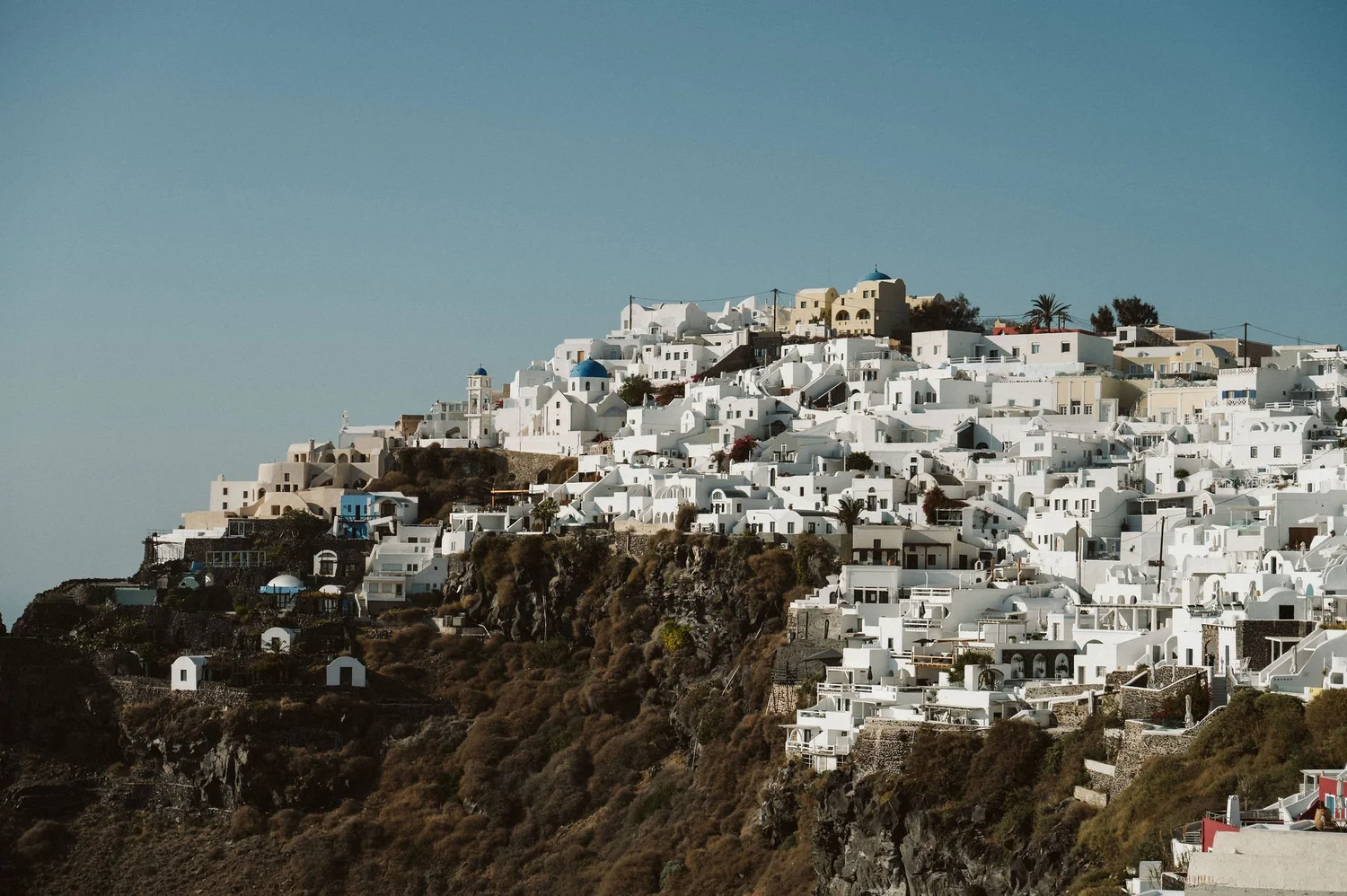 A hillside town with white buildings, some with blue domes, typical of Santorini, Greece, against a clear blue sky.