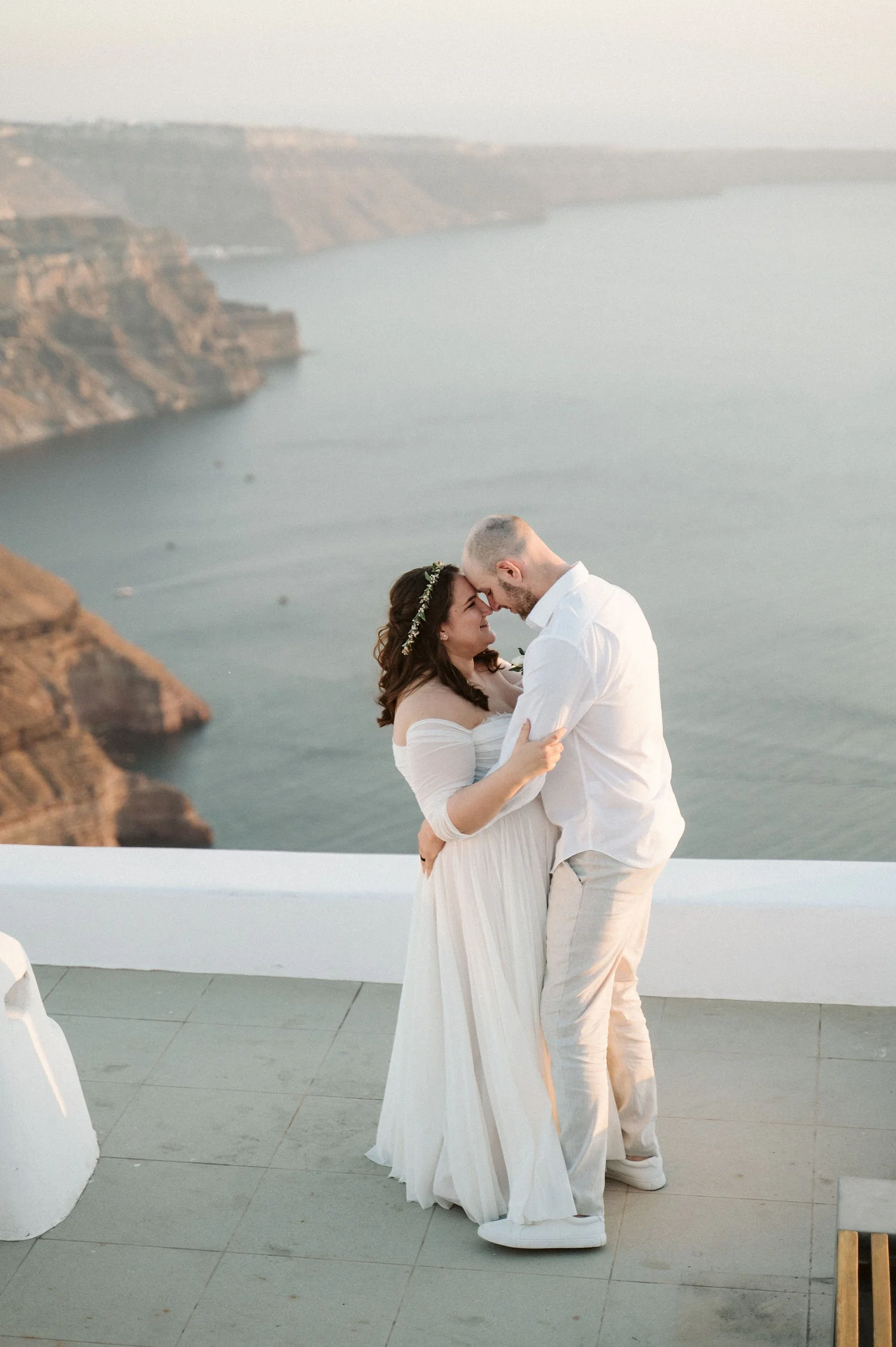 A couple dressed in wedding attire, sharing an intimate moment with foreheads touching against a scenic coastal backdrop.
