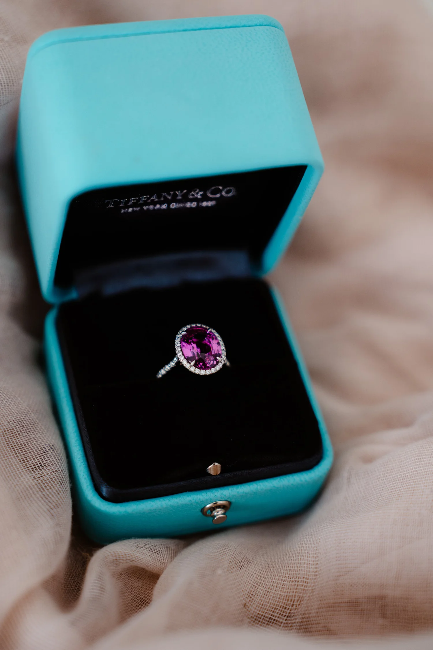 A jewelry ring featuring a large pink gemstone surrounded by smaller diamonds, displayed in a Tiffany & Co. jewelry box.