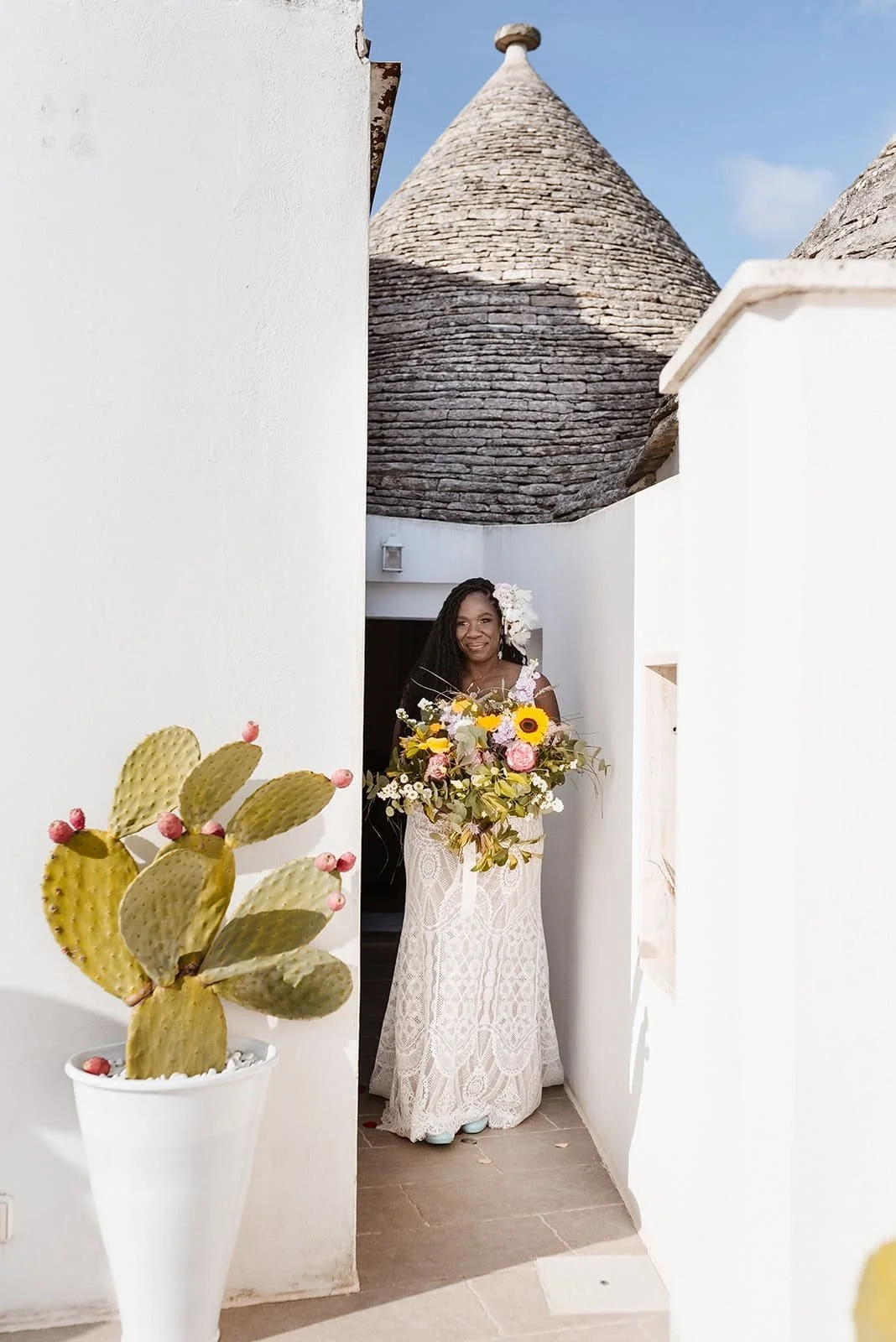 A woman in a lace wedding dress holding a bouquet of sunflowers, pink roses, and greenery standing in a narrow white-walled outdoor space with traditional conical stone roofs in the background and a potted prickly pear cactus in the foreground.