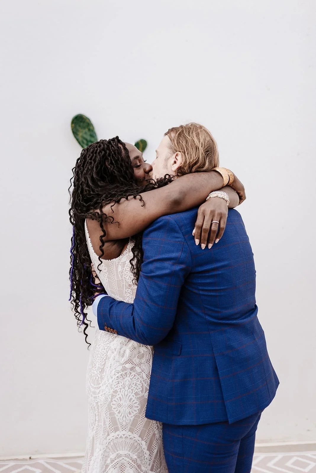 A couple embracing and kissing, the woman wearing a white lace dress and the man in a blue suit, against a plain white wall with a small green plant in the background.