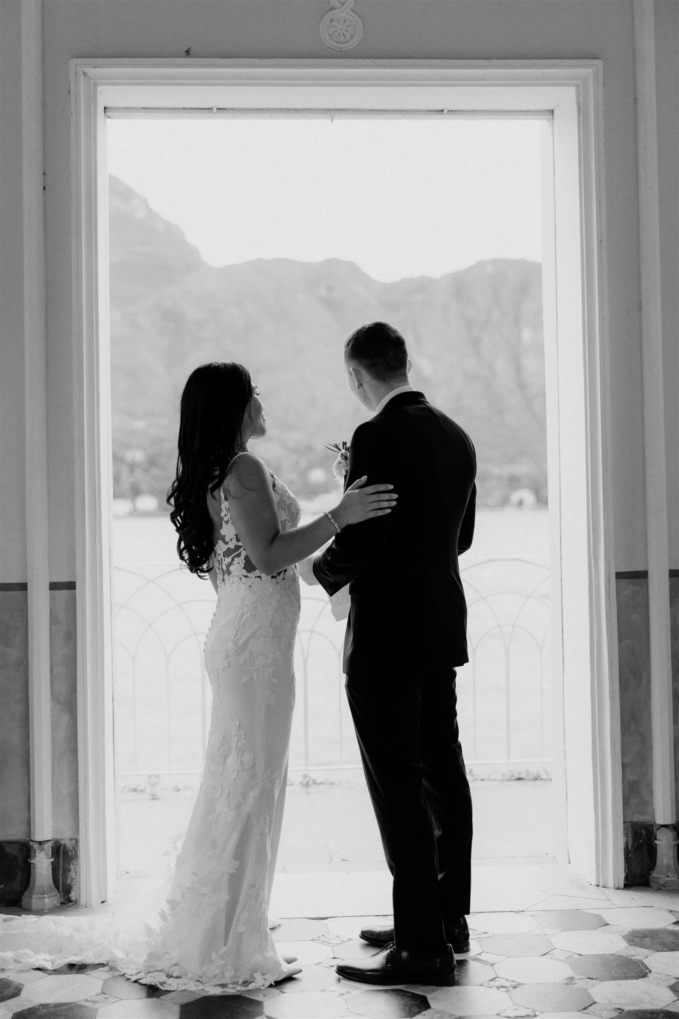 A black and white photo of a bride and groom facing each other, standing in front of a large window with mountains visible outside. The bride is wearing a lace wedding dress and the groom is in a suit. They are holding hands and looking at each other