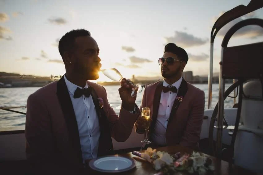 Two men dressed in tuxedos with pink jackets, black bow ties, and white shirts, are seated on a boat at sunset. One man is drinking from a wine glass, while the other is holding a glass of champagne. There is a table with a flower arrangement and an