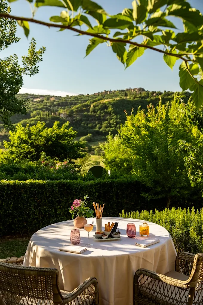 An outdoor dining table set for a meal, with a white tablecloth, glasses, bowls, and utensils, surrounded by greenery and rolling hills in the background.