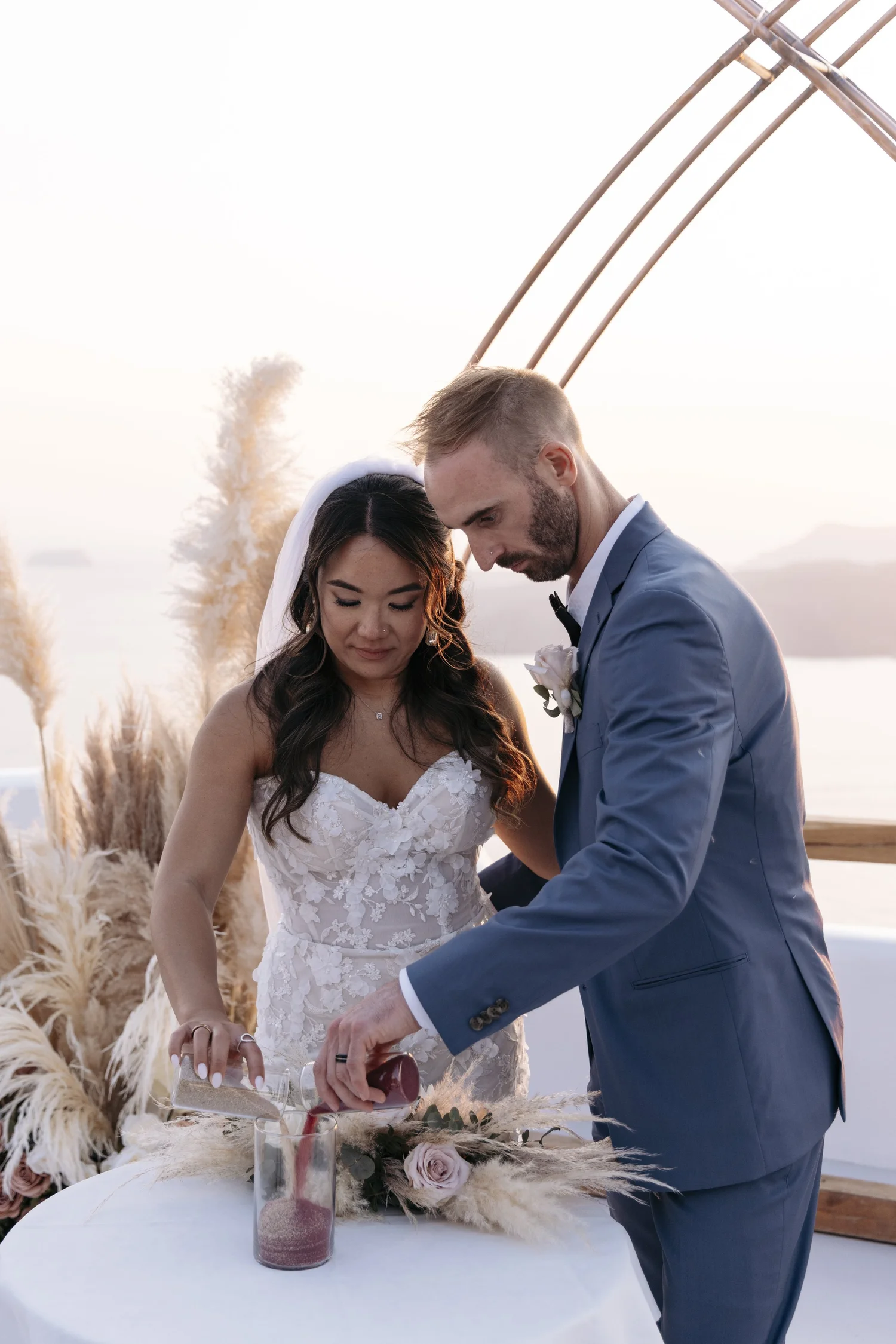 A bride and groom at a wedding ceremony signing a document outdoors at sunset, with a table decorated with flowers and pampas grass, and a scenic water view in the background.
