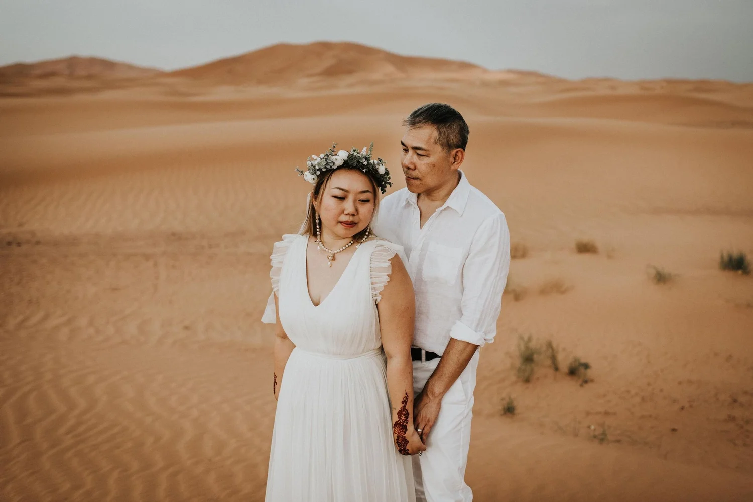 A couple dressed in white standing in a desert landscape with rolling sand dunes in the background.