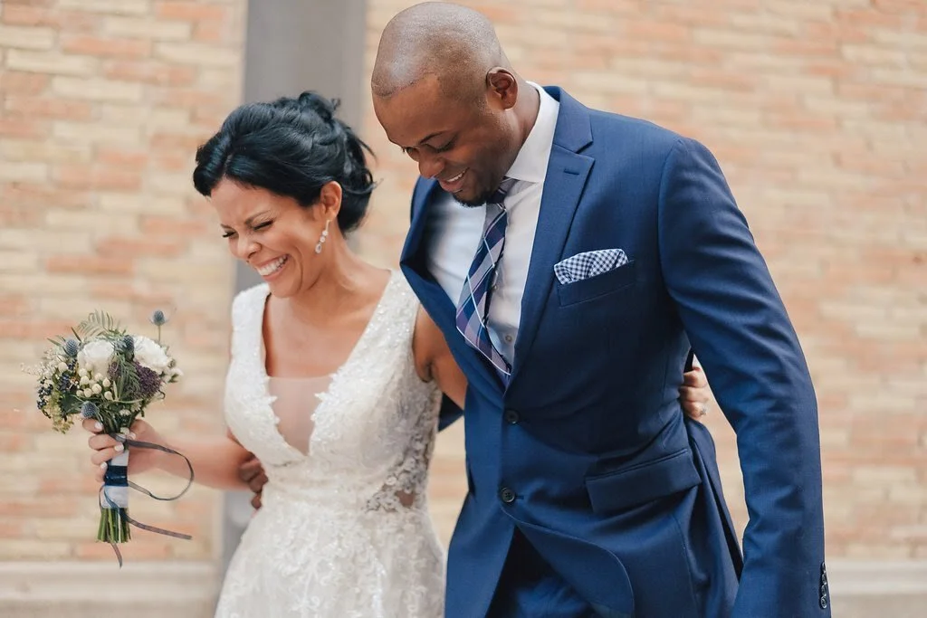 A bride in a white lace wedding dress holding a bouquet, standing beside a groomsman in a navy blue suit, both smiling and leaning towards each other.
