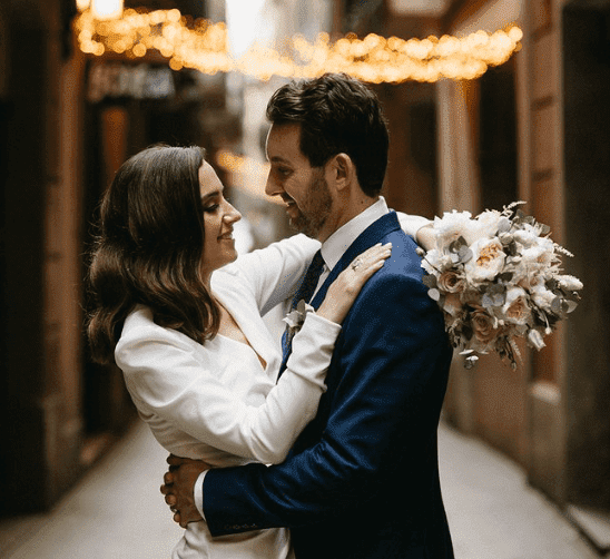 A couple embracing at night, with string lights overhead, on a city street decorated for an event, possibly a wedding, with the woman holding a bouquet of flowers.