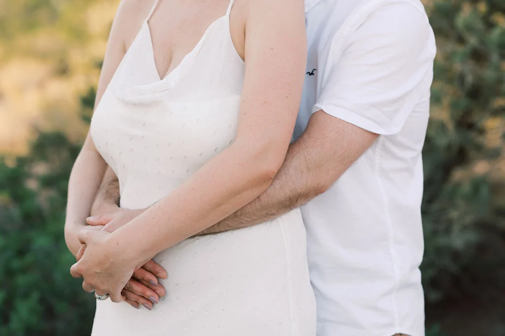 A couple holding hands, with the woman in a white dress and the man in a white shirt, outdoors with blurred greenery in the background.