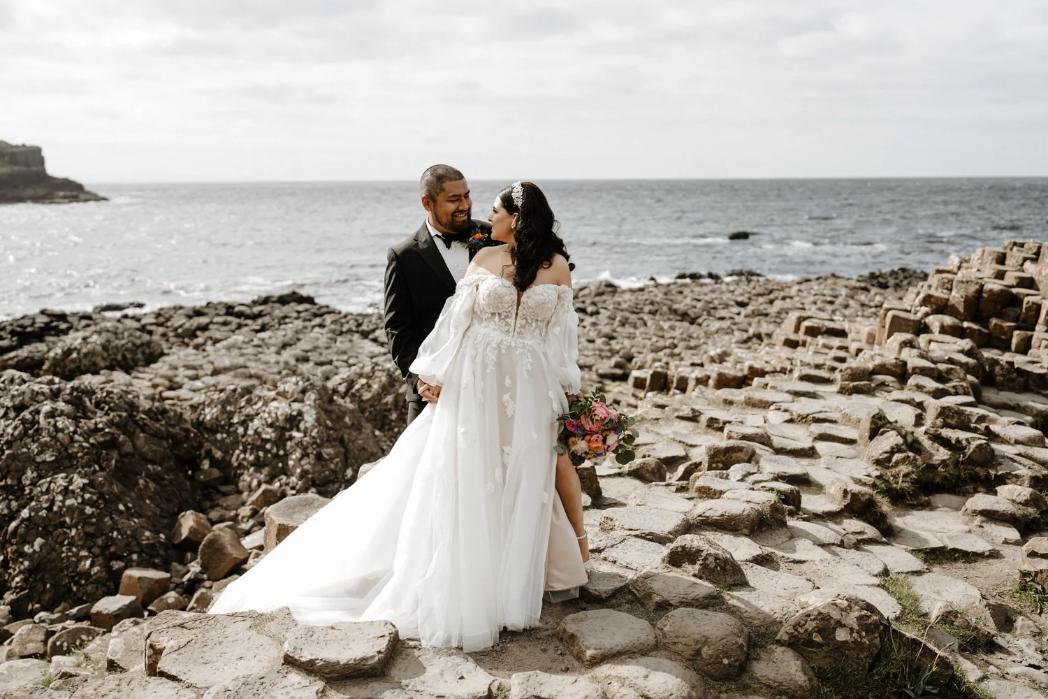 A bride and groom standing on a rocky beach, facing each other and smiling, with the ocean and cloudy sky in the background. The bride is wearing a white off-shoulder wedding gown and holding a colorful bouquet, while the groom is in a black tuxedo.