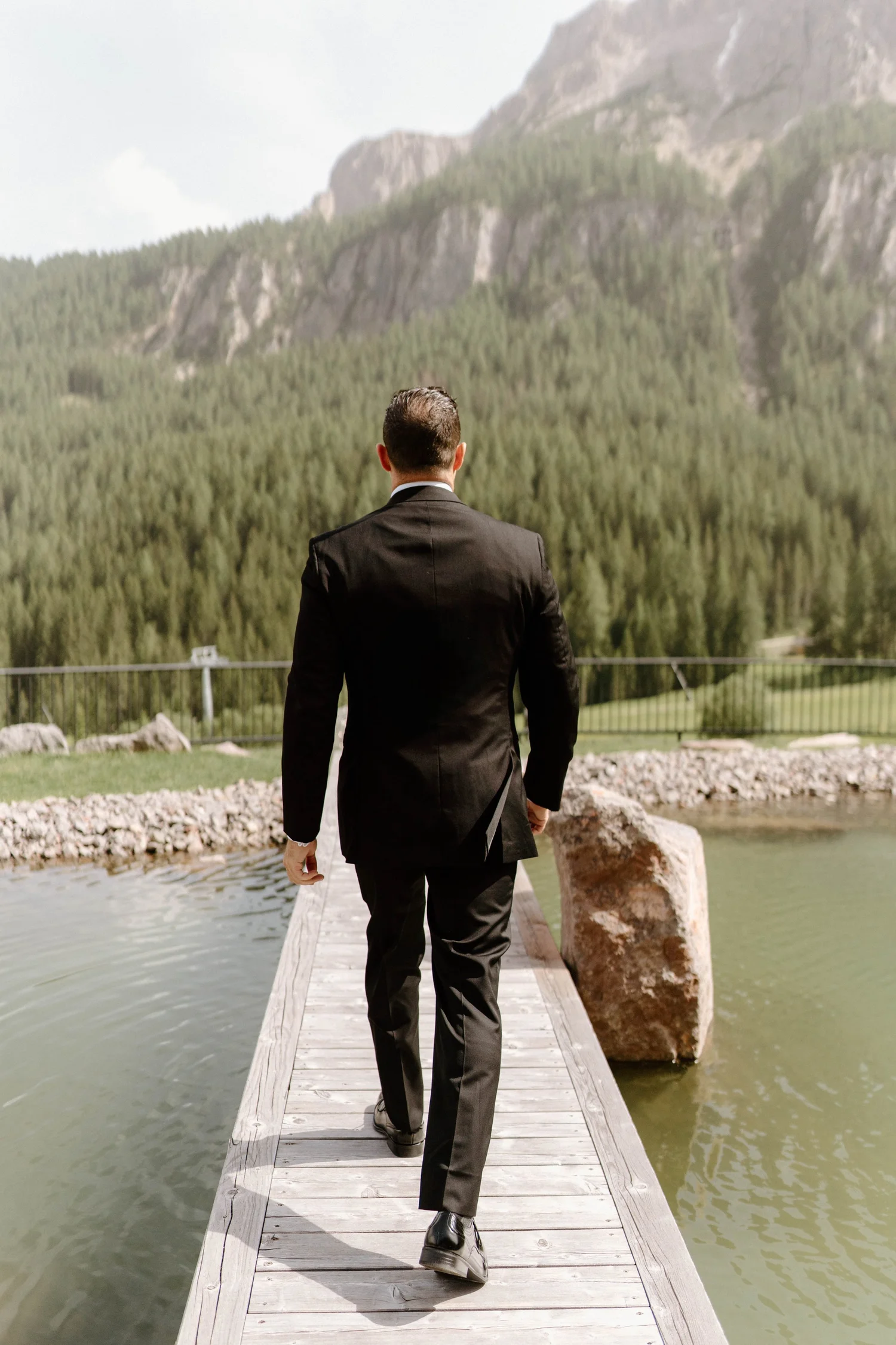 A man in a black suit walking on a wooden dock over water, with large rocks and a mountain covered in trees in the background.