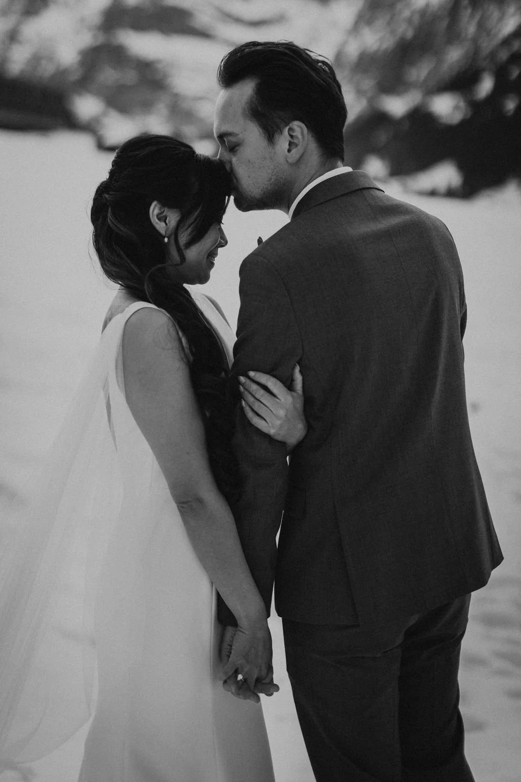 A black and white photo of a couple on their wedding day, with the man kissing the woman's forehead and holding her hand, outdoors near water.