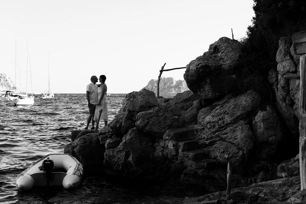 A black and white photo of a couple standing on rocks near the ocean, holding hands and facing each other, with boats in the background and a large rock formation on the right.