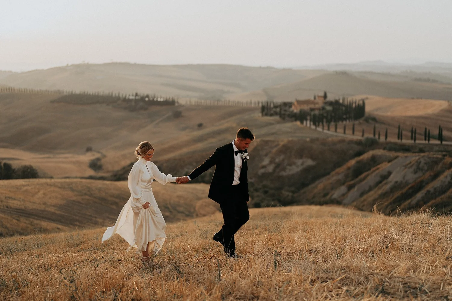A bride and groom holding hands and walking through a grassy field in a scenic landscape with rolling hills and scattered trees.